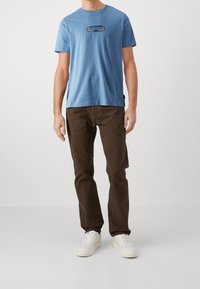 Jasnoniebieska koszulka bawełniana z okrągłym dekoltem i logo na klatce piersiowej; w połączeniu z brązowymi spodniami z kordurowej tkaniny i białymi trampkami.