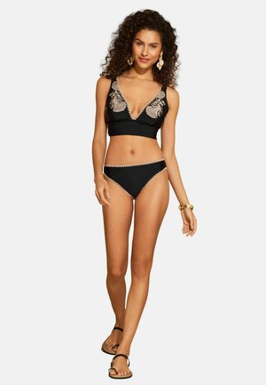 Čierny bikini set s braletom do V výstrihu zdobeným béžovou výšivkou na okrajoch, doplnený o zodpovedajúce bikini nohavičky a čierne sandále.