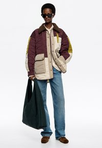 Chaqueta acolchada en burdeos, crema y oliva, con un cuello de faux fur marrón, bolsillos frontales y botones. Combinada con jeans de mezclilla y un bolso verde.
