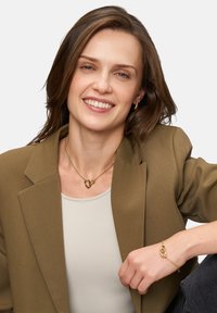 Goldene Halskette mit verflochtenem Herzdesign und passendem goldenen Armband, getragen mit einem beigen Oberteil und olivgrüner Blazer.