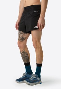 Černé sportovní šortky s elastickým pasem, logo na boku. Model má na sobě modrou obuv se šedými detaily a černo-turkvazové ponožky.