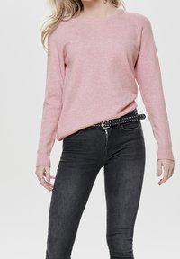 Hellrosa gestrickter Pullover mit rundem Halsausschnitt, gerippten Bündchen und Saum. Kombination mit dunkelgrauen, eng anliegenden Jeans und einem schwarzen Nieten-Gürtel.