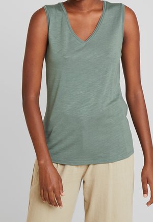 Persona con una camiseta sin mangas de color verde salvia con escote en V y pantalones beige claro, de pie con los brazos relajados a los lados.