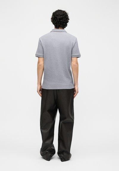 Άνδρας με σγουρά μαλλιά, κοιτάζοντας προς τα πίσω, φορά γκρι πόλο με κοντά μανίκια και φαρδιά μαύρη παντελόνια.