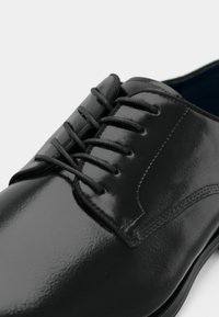 Pantofii eleganți din piele lăcuită neagră, cu șireturi închise, cusături detaliate și vârf rotund, pe un fundal alb.