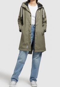 Oliven grøn vandtæt jakke med hætte, featuring sort foring, hvide snoretræk og trykknapper. Båret over lyseblå jeans og hvide sneakers.