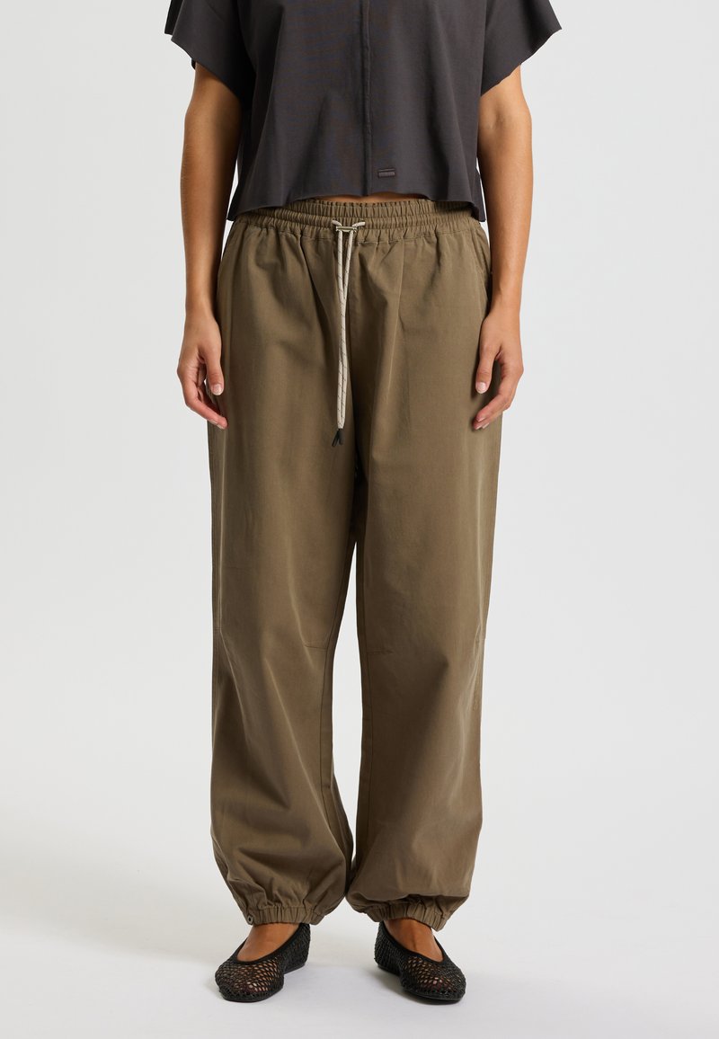 Housut, joissa on löysä istuvuus, khaki-väriset, joustava vyötärö ja nauhakuja, säädettävät lahkeensuut. Yhdistetty tumman väriseen lyhythihaiseen toppiin ja mustiin kengiin.