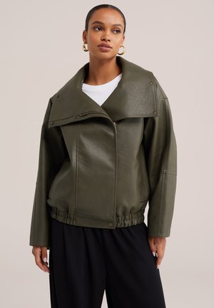 Femme portant une veste en cuir vert foncé oversize avec un grand col, une chemise blanche, un pantalon noir et des boucles d’oreilles anneaux dorés sur un fond uni.