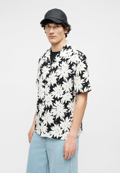 Jeune homme portant une casquette noire, une chemise à manches courtes noire et blanche à motifs floraux, et un jean bleu clair, debout devant un fond blanc uni.