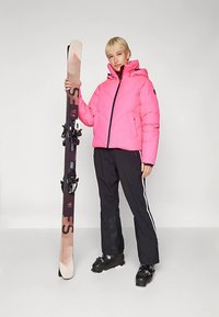 Ροζ μπουφάν τύπου puffer με διαγώνια ραφή, μαύροι σκι παντελόνια με λευκές πλαϊνές ρίγες και μαύρα μποτάκια για σκι. Κρατά ένα καφέ σκι με δέστρες.