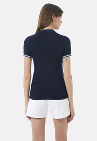 Polo shirt blu navy con un design aderente, maniche corte e bordature bianche sul colletto e sui bordi delle maniche. Abbinato a pantaloni bianchi.