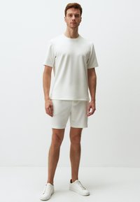 T-shirt bianco a maniche corte e pantaloni corti abbinati realizzati in tessuto morbido. Il modello indossa sneakers bianche, mentre è in piedi contro uno sfondo semplice.