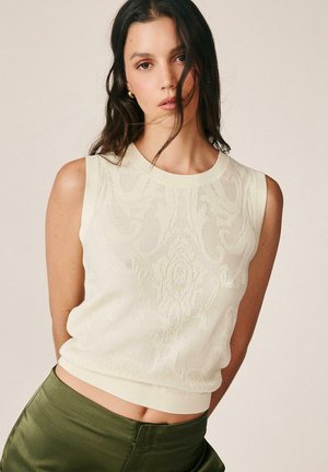 Mujer con cabello largo y oscuro que lleva un top de punto sin mangas color crema y pantalones verde oliva sobre un fondo liso.