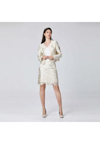 Femme portant une veste en brocart floral crème et une jupe assortie, un top blanc et des talons beiges, se tenant devant un fond gris clair uni.