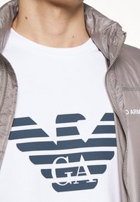 Ljusgrå jacka med dragkedja, lagd över en vit t-shirt med en blårandig logotyp. Slätt tyg och rena linjer.