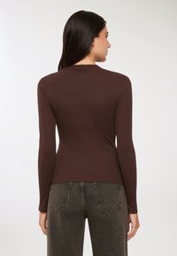 Kobieta z długimi ciemnymi włosami, ubrana w dopasowaną brązową bluzkę z długim rękawem i wyblakłe czarne dżinsy, pokazana od tyłu na białym tle.