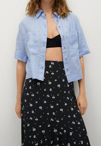 Camicia azzurra a maniche corte con bottoni, dotata di due tasche sul petto, abbinata a una gonna nera con piccoli motivi floreali blu.