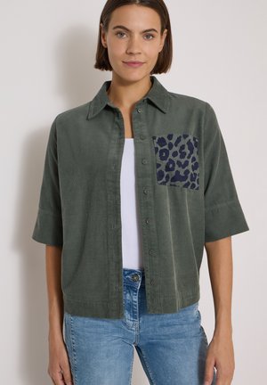 Femme portant une chemise en velours côtelé verte avec une poche poitrine imprimée léopard bleu foncé, un top blanc en dessous et un jean bleu clair.