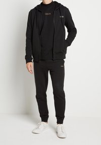 Zwarte zip-up hoodie met trekkoorden over een zwart T-shirt met korte mouwen en zwarte joggingbroeken. Gouden merkaccenten op het shirt en de broeken. Witte sneakers.