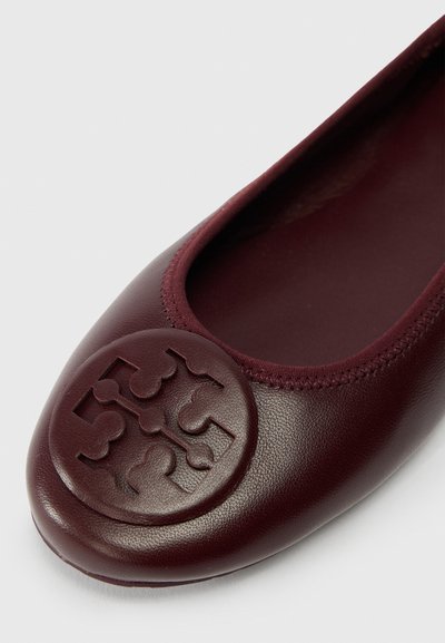 Close-up of a burgundy leather ballet flat with a round, embossed decorative emblem on the toe and stitched trim around the opening.