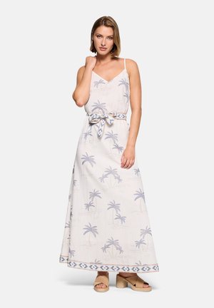 Mujer con vestido largo blanco con estampado de palmeras azules y cinturón atado, de pie con la mano cerca del rostro, sandalias de tacón marrón claro.