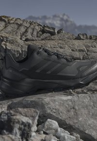 Chaussure de randonnée Adidas Terrex noire posée sur un terrain rocheux avec des montagnes floues en arrière-plan sous un ciel dégagé.