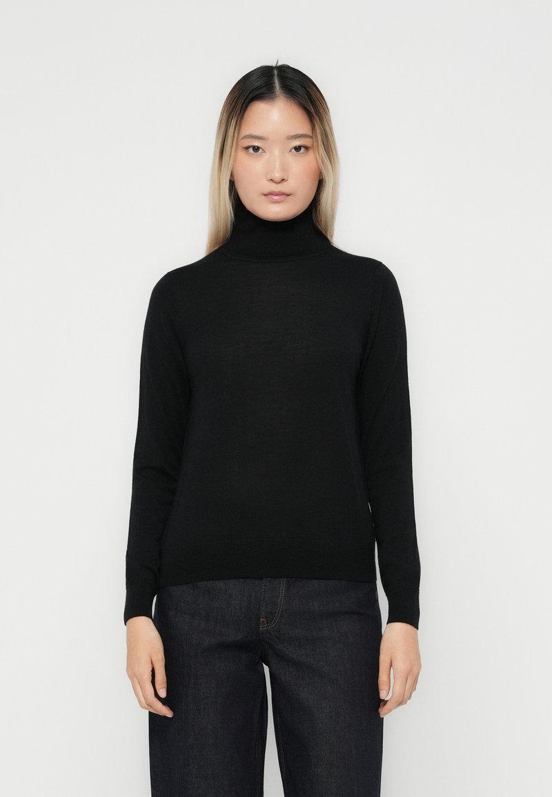 Pull à col roulé noir en maille fine. Manches longues, design ajusté et style minimaliste. Porté avec un pantalon en denim sombre.