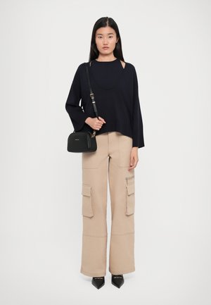 Femme avec des cheveux noirs raides portant un pull noir ample, un pantalon cargo beige à jambe large, des chaussures noires à bout pointu et portant un sac en bandoulière noir.