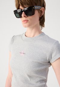 Crne oversized sunčane naočale s sjajnom završnom obradom, nosi se s svijetlosivim, rebrastim majicom s kratkim rukavima i okruglim izrezom koja ima mali ružičasti logo Calvin Klein.