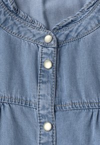 Tissu en denim bleu clair avec trois boutons-pression métalliques et coutures visibles, montrant une partie de la fermeture avant d’un vêtement.