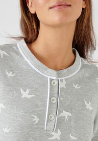 Camisa de algodón gris con cuello redondo, ribete blanco, tapeta de botones y un bolsillo, con un patrón de pájaros en toda la prenda.