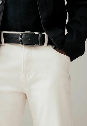 Black leather belt with a silver metal buckle paired with light cream-colored pants and a black textured top. Focus on the waist area.