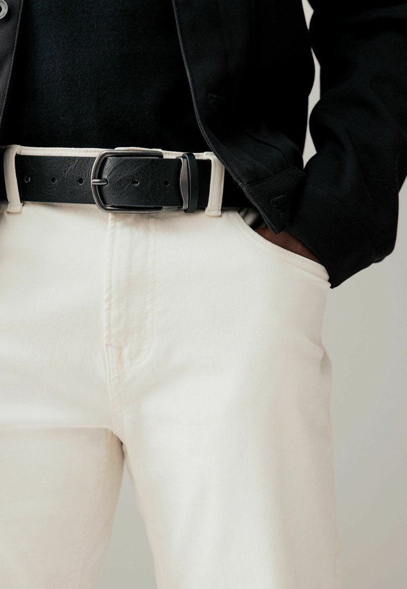 Ceinture en cuir noir avec une boucle en métal argenté, associée à un pantalon crème clair et un haut texturé noir. Accent sur la taille.