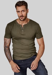 Olive green short-sleeve henley shirt with three buttons, fitted design, soft texture. Model wearing dark blue jeans, displayed against a gray background.