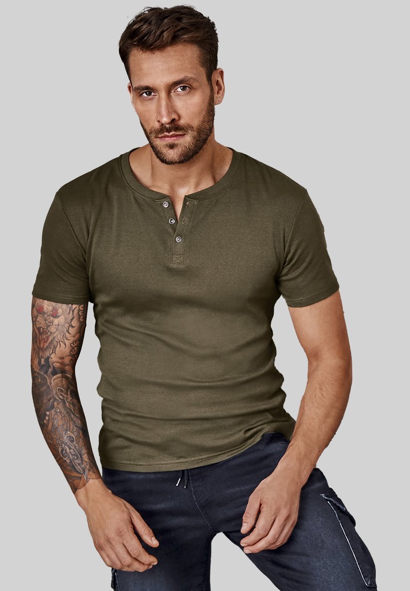 Olive green short-sleeve henley shirt with three buttons, fitted design, soft texture. Model wearing dark blue jeans, displayed against a gray background.