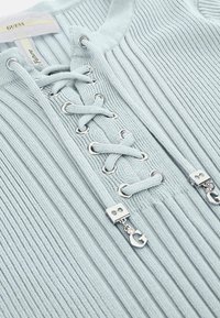 Šviesiai mėlynas raukšlėtas megztinis su juostelėmis priekyje, kuriame yra sidabrinės detalės ir dekoratyvūs metaliniai akcentai ant juostelių galų.