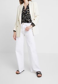 Kremowa dwurządowa marynarka na czarnej kwiatowej bluzce, zestawiona z białymi spodniami typu wide-leg i czarnymi klapkami z klamrami.