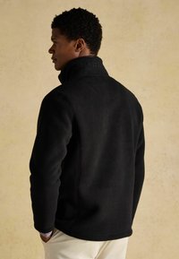 Giacca in pile nera con colletto alto, caratterizzata da una finitura testurizzata e un logo in rilievo sul colletto. Design semplice con maniche lunghe.