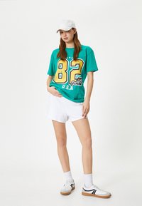 Større teal t-shirt med gult nummer "82" og teksten "U.S.A.", parret med hvide shorts og hvide sneakers med sorte detaljer.