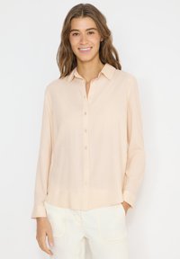 Femme aux cheveux bruns ondulés portant une chemise beige clair à manches longues boutonnée et un pantalon blanc, souriant devant un fond uni.