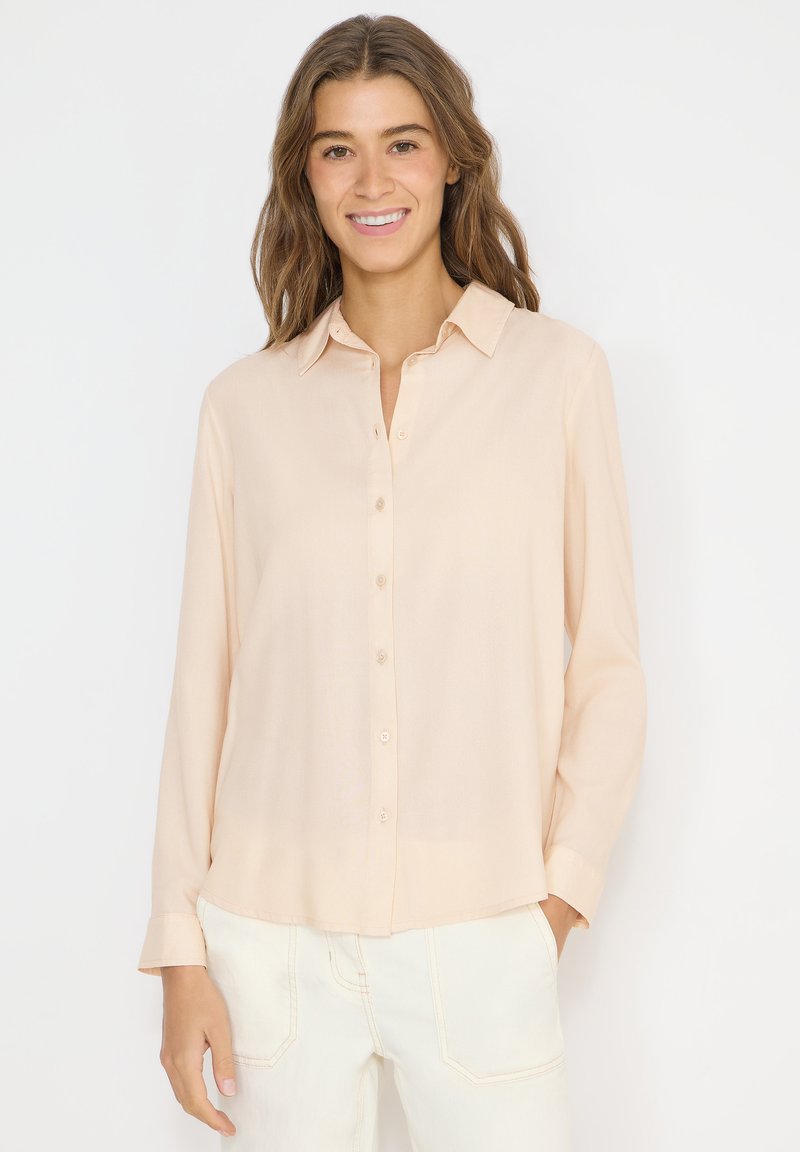Femme aux cheveux bruns ondulés portant une chemise beige clair à manches longues boutonnée et un pantalon blanc, souriant devant un fond uni.