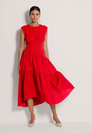 Rode jurk tot op de knie met een nauwsluitend lijfje en een uitlopende, gelaagde rok. Mouwloze cap sleeves en een gestructureerde stof, gecombineerd met lichtgekleurde schoenen.