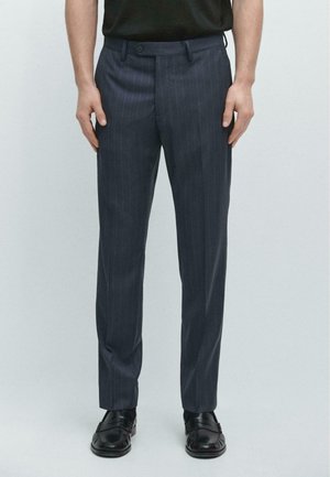 Mann trägt marineblaue Nadelstreifenhose, schwarzen Gürtel, schwarze Loafer und ein in die Hose gestecktes schwarzes Hemd, steht vor einem einfarbigen hellen Hintergrund.