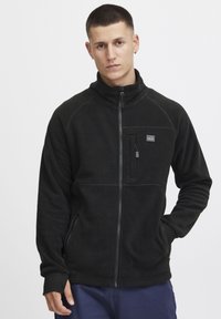 Schwarze Fleecejacke mit hohem Kragen und Reißverschluss. Hat eine Brusttasche auf der linken Seite, seitliche Taschen und ein strukturiertes Gewebe.