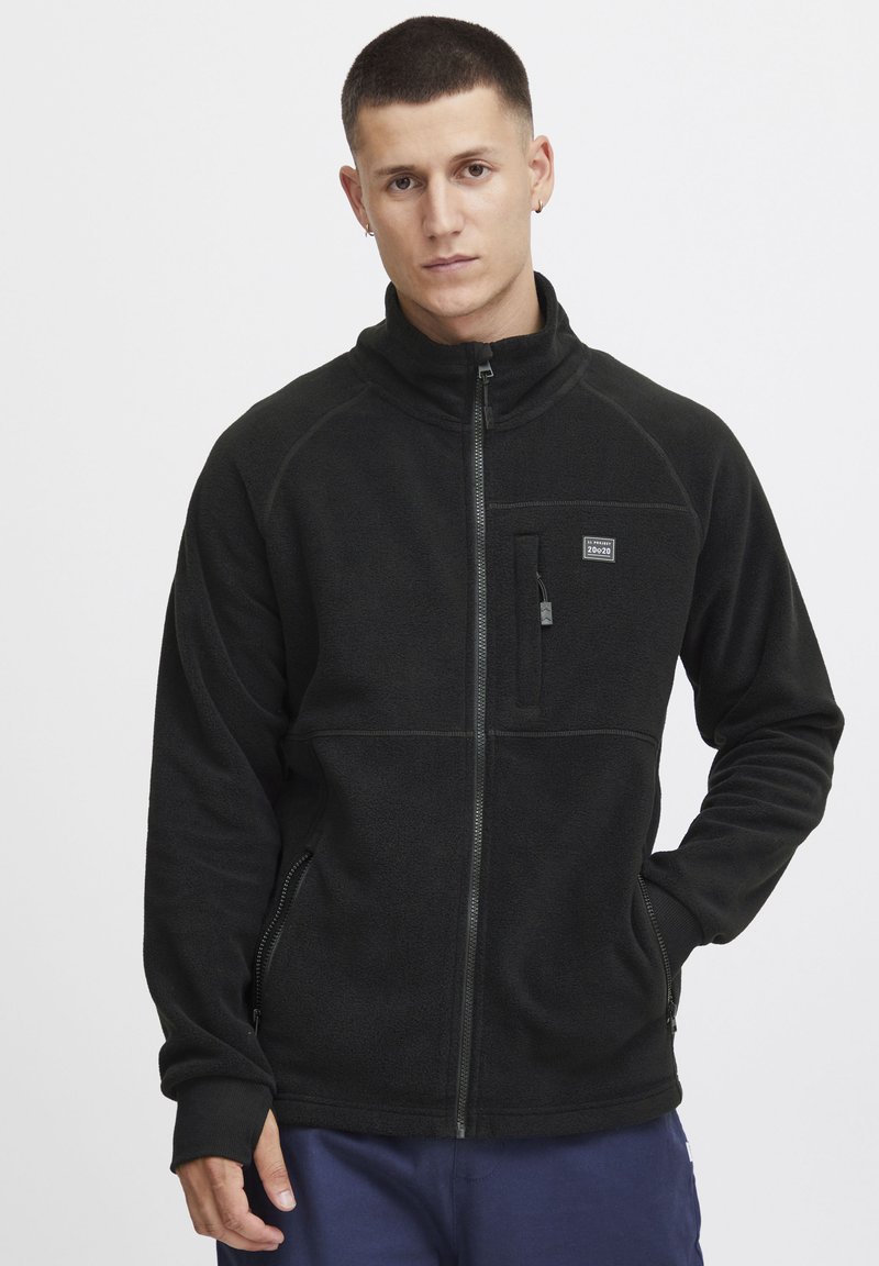 Schwarze Fleecejacke mit hohem Kragen und Reißverschluss. Hat eine Brusttasche auf der linken Seite, seitliche Taschen und ein strukturiertes Gewebe.