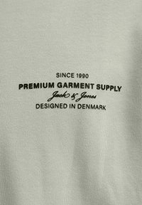 Zeleni majica iz blaga s črnim besedilom: "OD LETA 1990 PREMIUM GARMENT SUPPLY Jack & Jones NAČRTOVANO NA DANSKEM". Gladka, teksturirana površina.