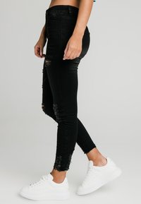 Svarta, slitna skinny jeans med fransar vid fållen och revor vid knäna, tillsammans med vita sneakers. Material: denim.
