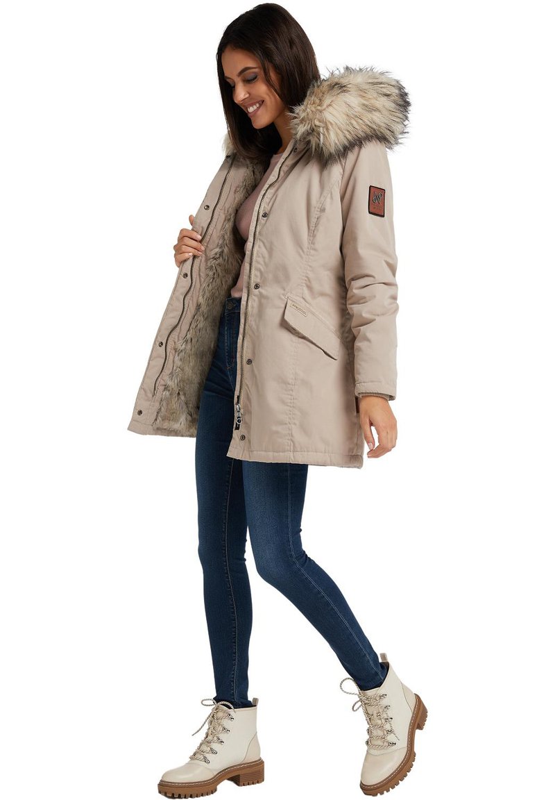 Beige parka with faux fur hood and lining, zip and snap closure, side pockets, and brown leather accents. Paired with blue jeans and boots.