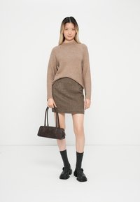 Beige Strickpullover, karierten braunen Rock, schwarze Knöchelsocken, klobige Schuhe und eine dunkle Handtasche mit doppelten Griffen.