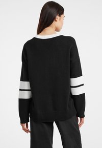 Pull en tricot noir avec un large col, ourlet côtelé et rayures blanches contrastées sur chaque manche. Texture douce, coupe décontractée.